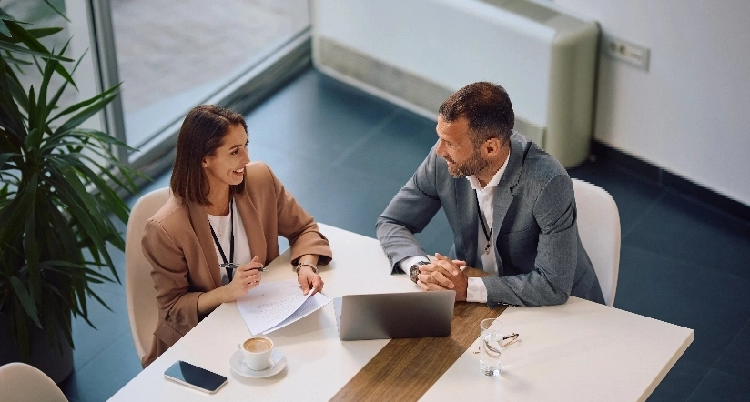 A man and woman sit at a table, focused on a laptop, engaged in discussion or collaboration.