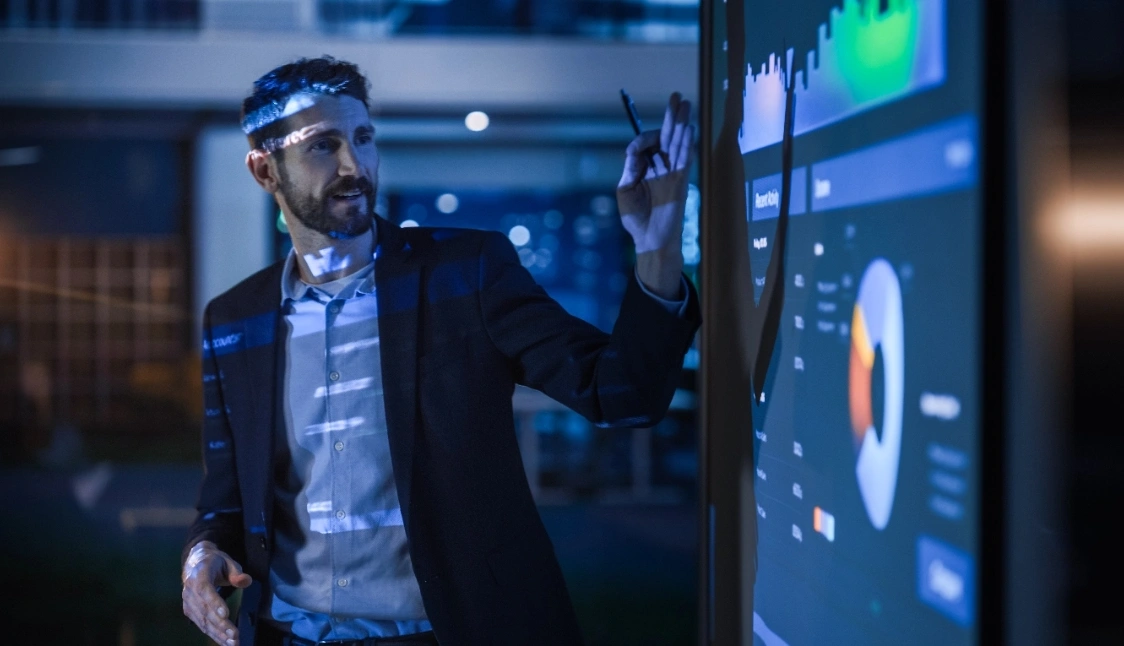A man in a suit points at a screen, likely presenting information to an audience