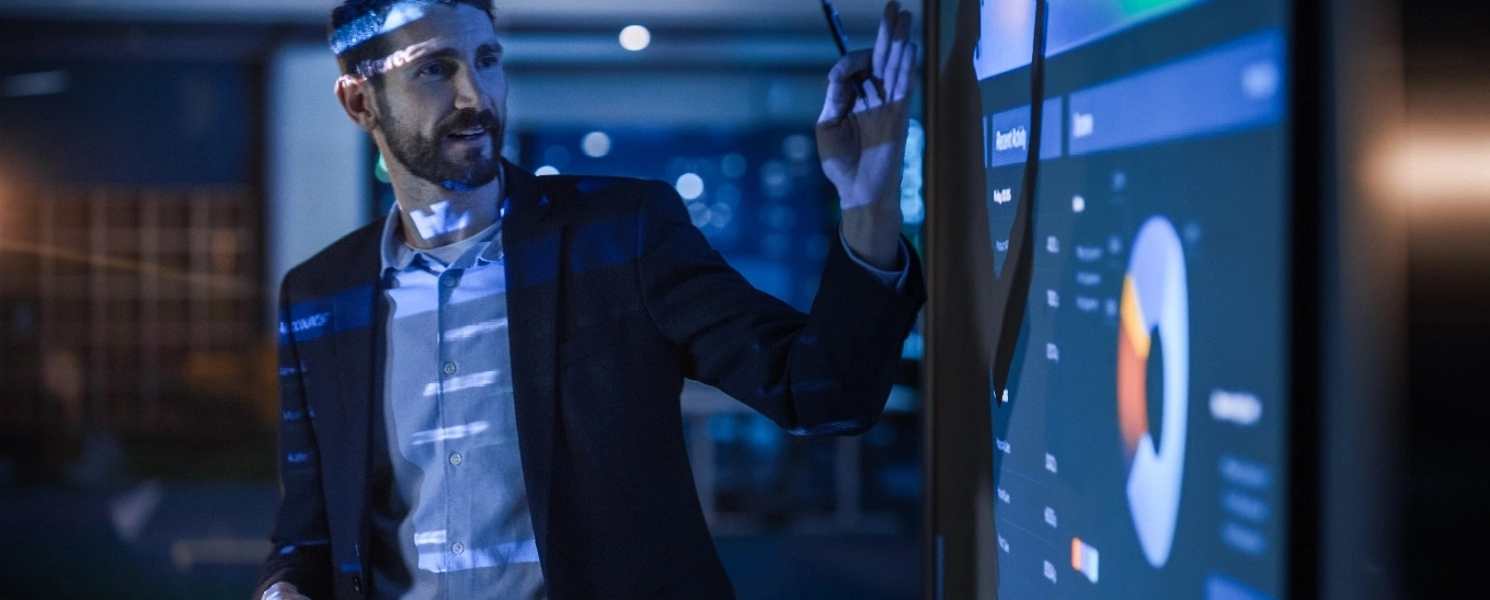 A man in a suit stands confidently in front of a large screen displaying data or visuals