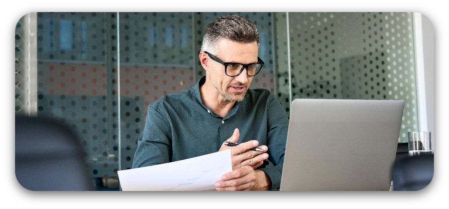 A man wearing glasses sits at a desk with a laptop open in front of him