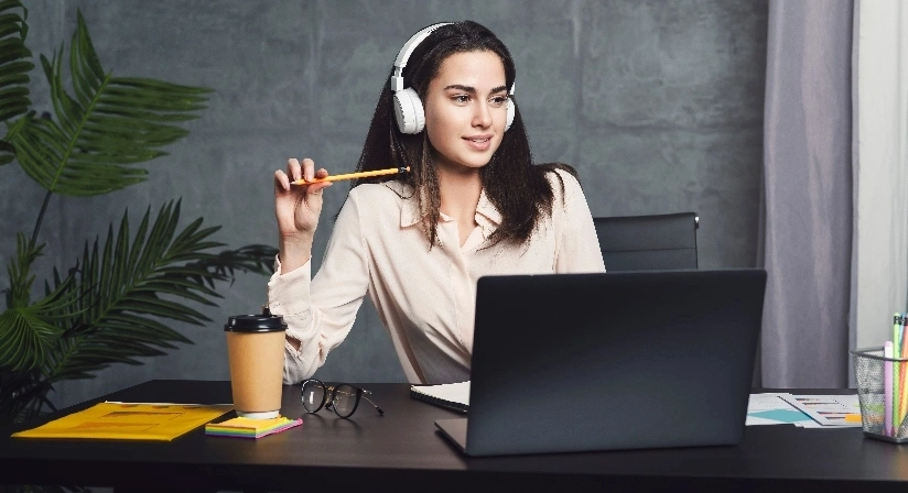 A woman with headphones is seated at a desk, engaged with her laptop, indicating a work or study environment