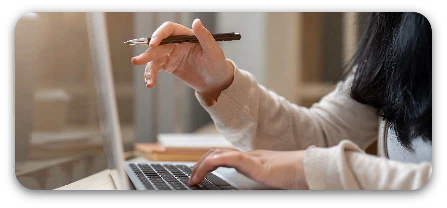 A woman writes with a pen while working on a laptop at a desk, focused on her task