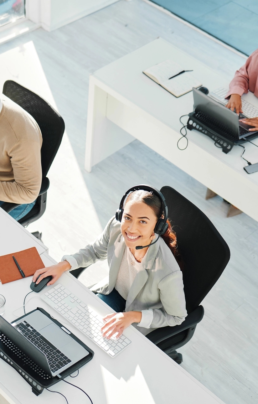 High-angle shot of a smiling woman wearing a headset while working at a desk with a keyboard, computer, and mouse, in an open office with other workers